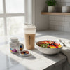 Supplement containers, a protein shaker, and a bowl of fruit on a kitchen counter.